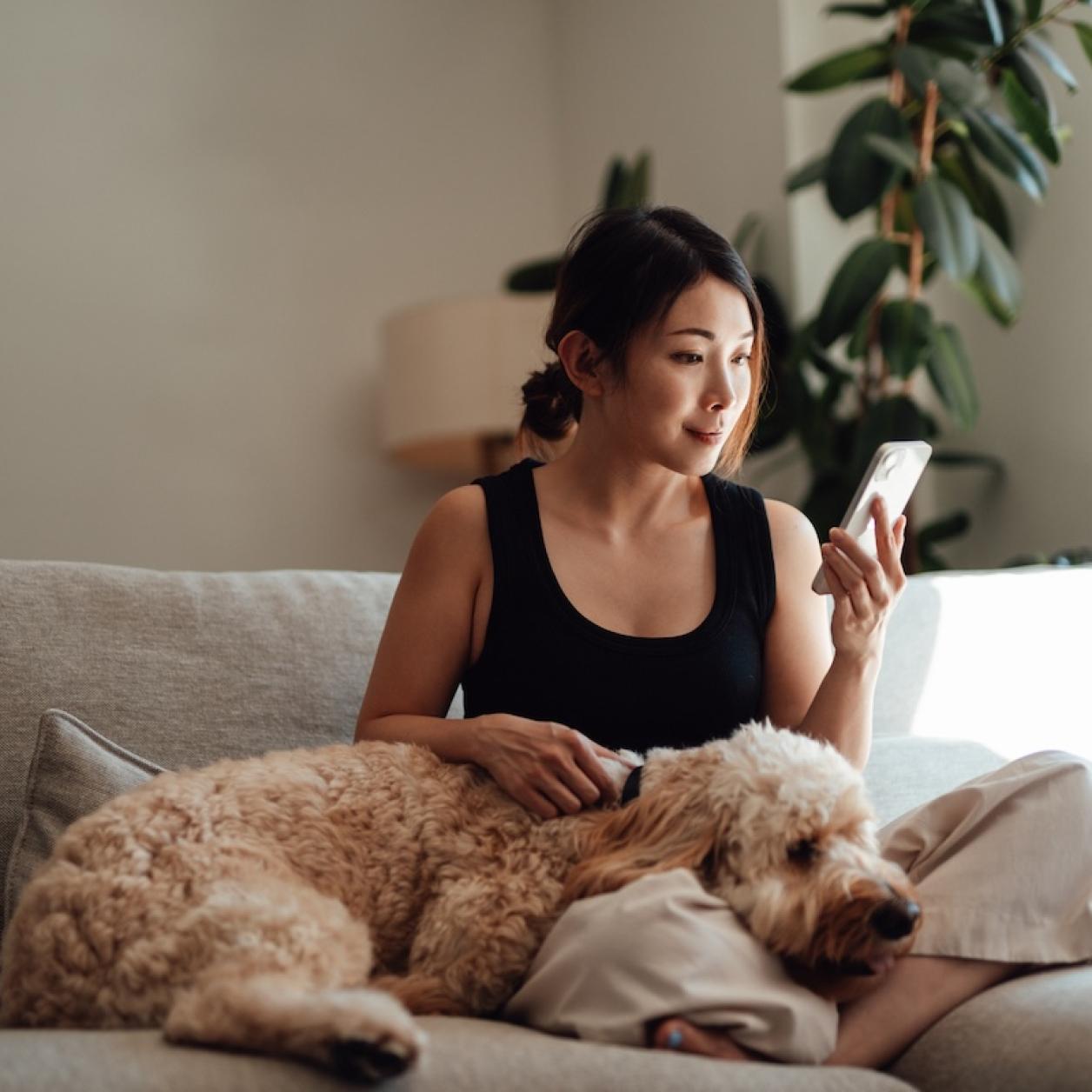 A woman sits on a couch with a dog and looks at her phone